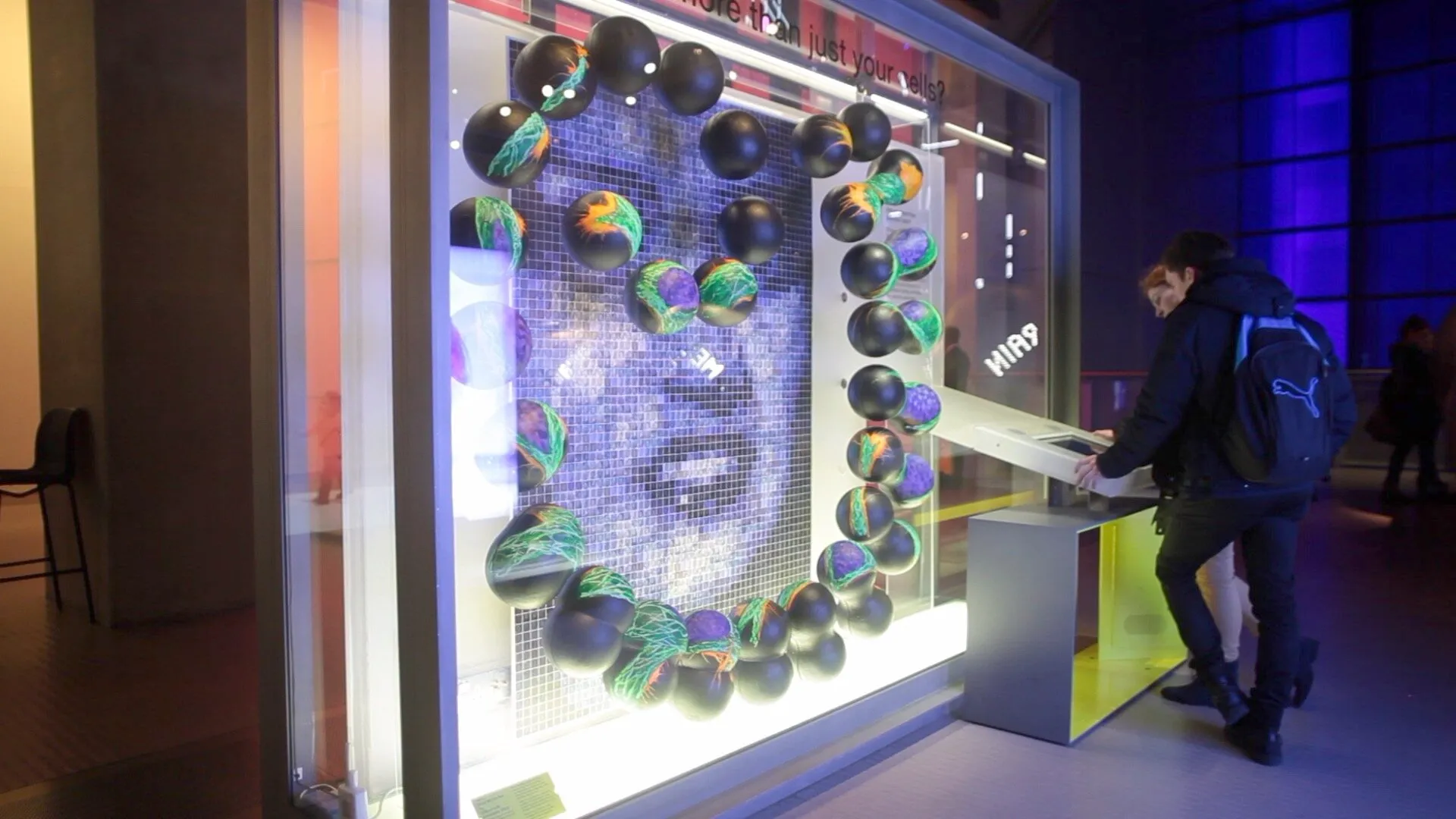 Wide view of the display case with the sphere sculptures and mosaic wall, with a visitor using an interactive touchscreen kiosk alongside