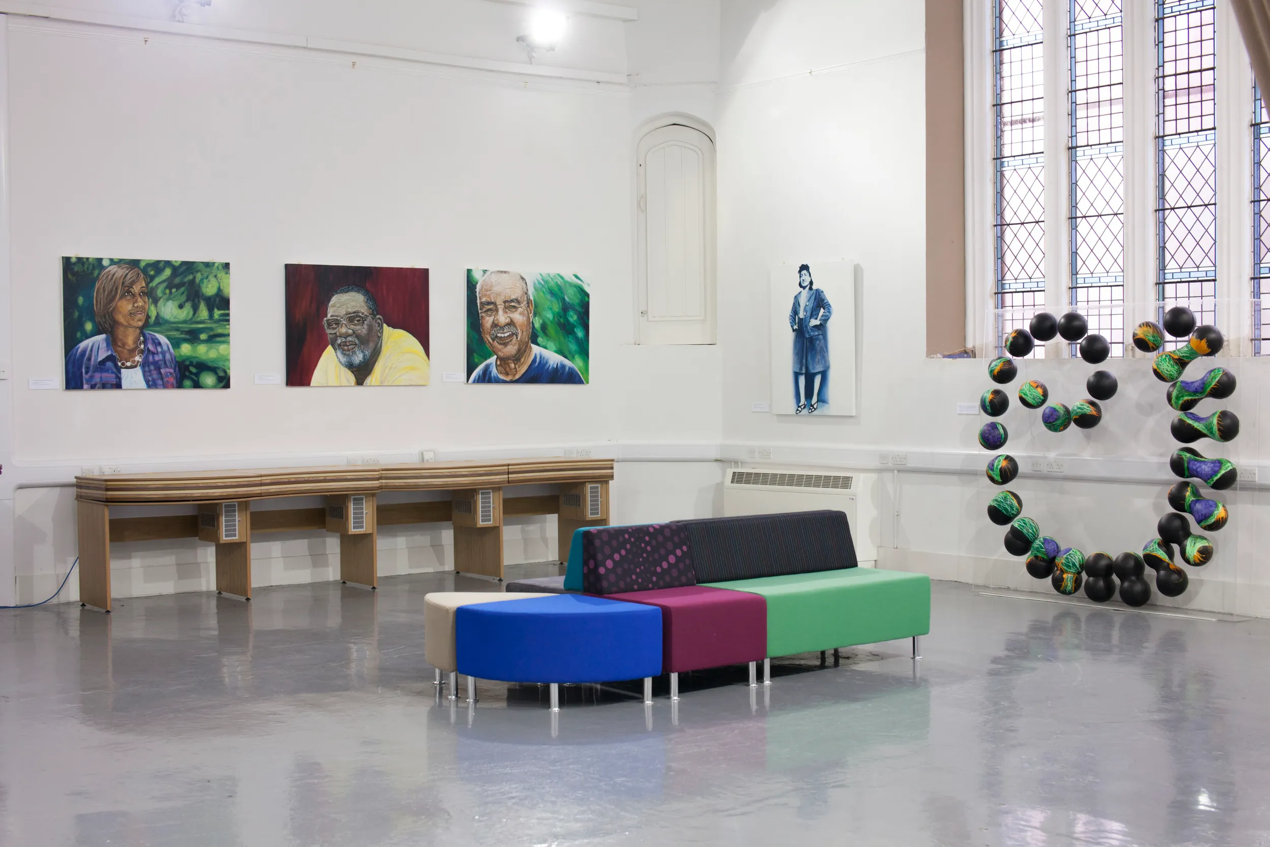 Gallery wall hung with painted portraits of three Lacks family members, a full-length portrait of Henrietta Lacks, and a sphere sculpture representing cells in the corner