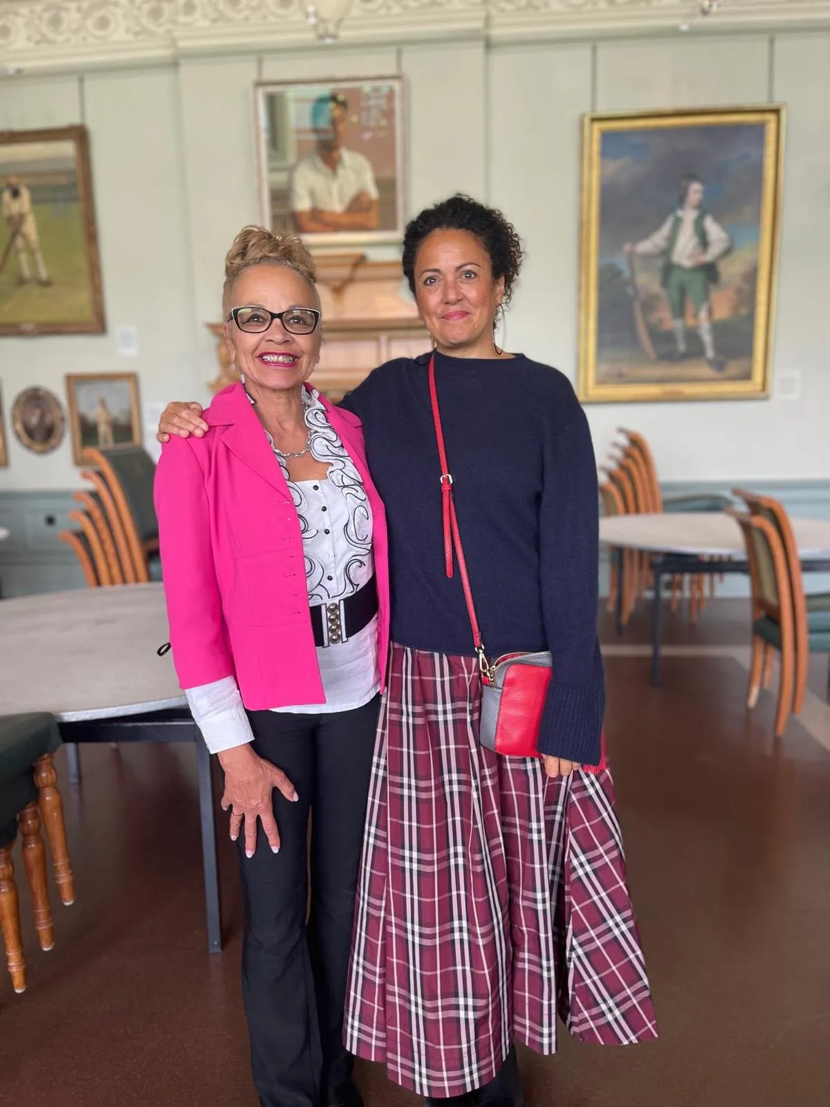 Anita Neil MBE and artist Helen Wilson Roe posing arm in arm in the MCC Museum at Lord's, with cricket portraits, including Helen's painting of Guy Bailey, hanging on the wall behind them