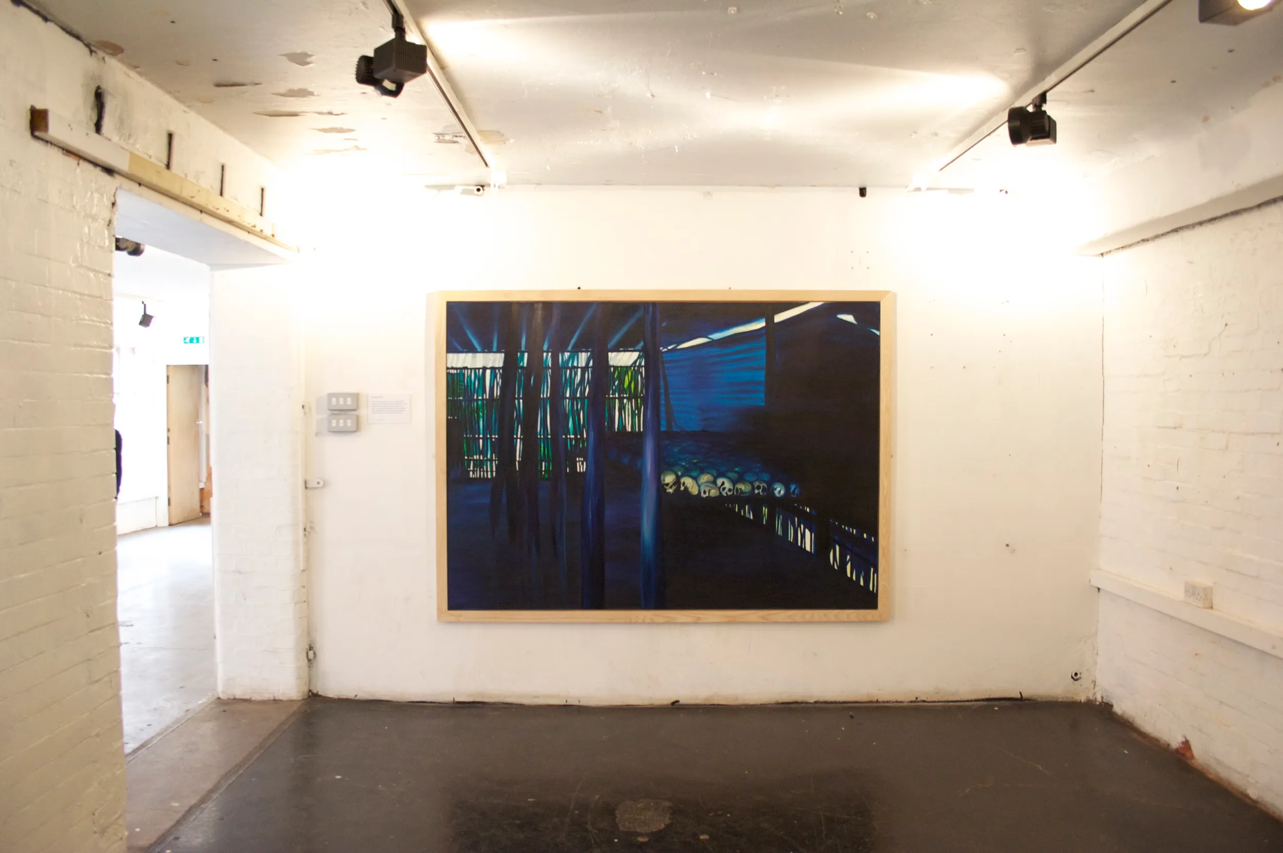Painting of a dimly lit genocide memorial interior, with rows of skulls displayed along a low wall and blue light streaming through slatted windows