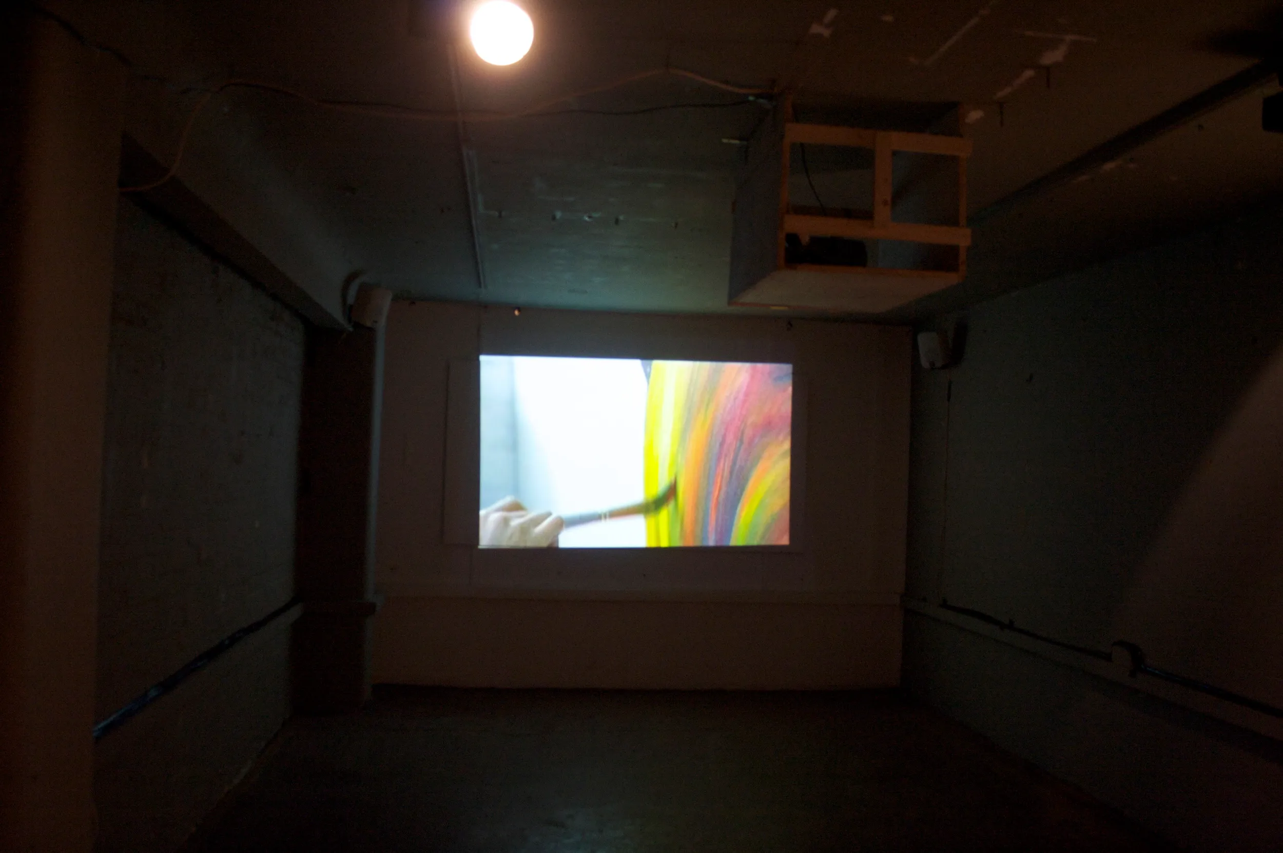Darkened gallery room with a video projection showing a gloved hand painting broad rainbow-coloured brushstrokes, from the documentary about the artist's work