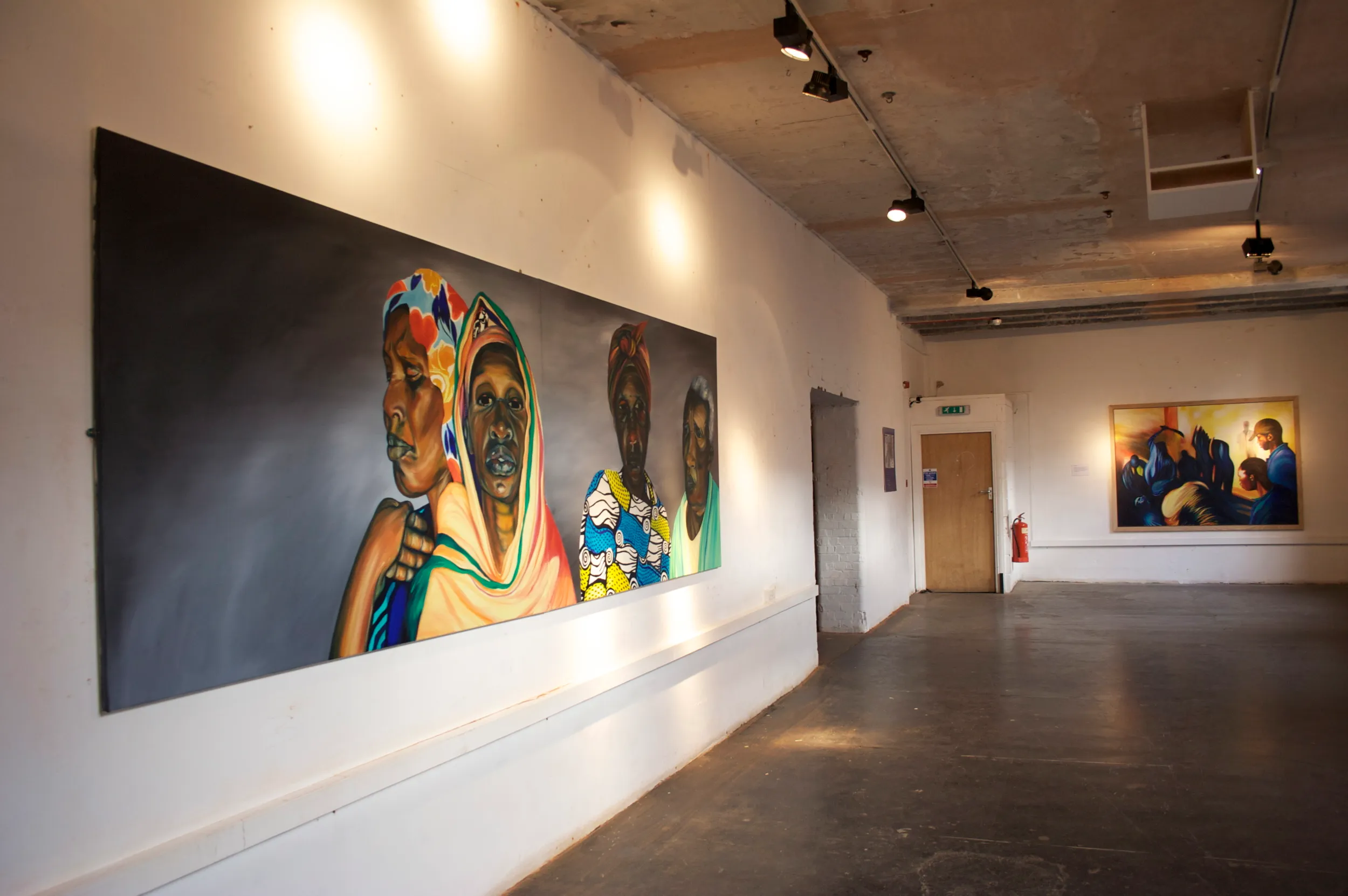 Gallery wall with a large painting of four Rwandan women in colourful headscarves and patterned clothing standing close together, and further along a smaller painting of dark bird-like forms and men against a golden background