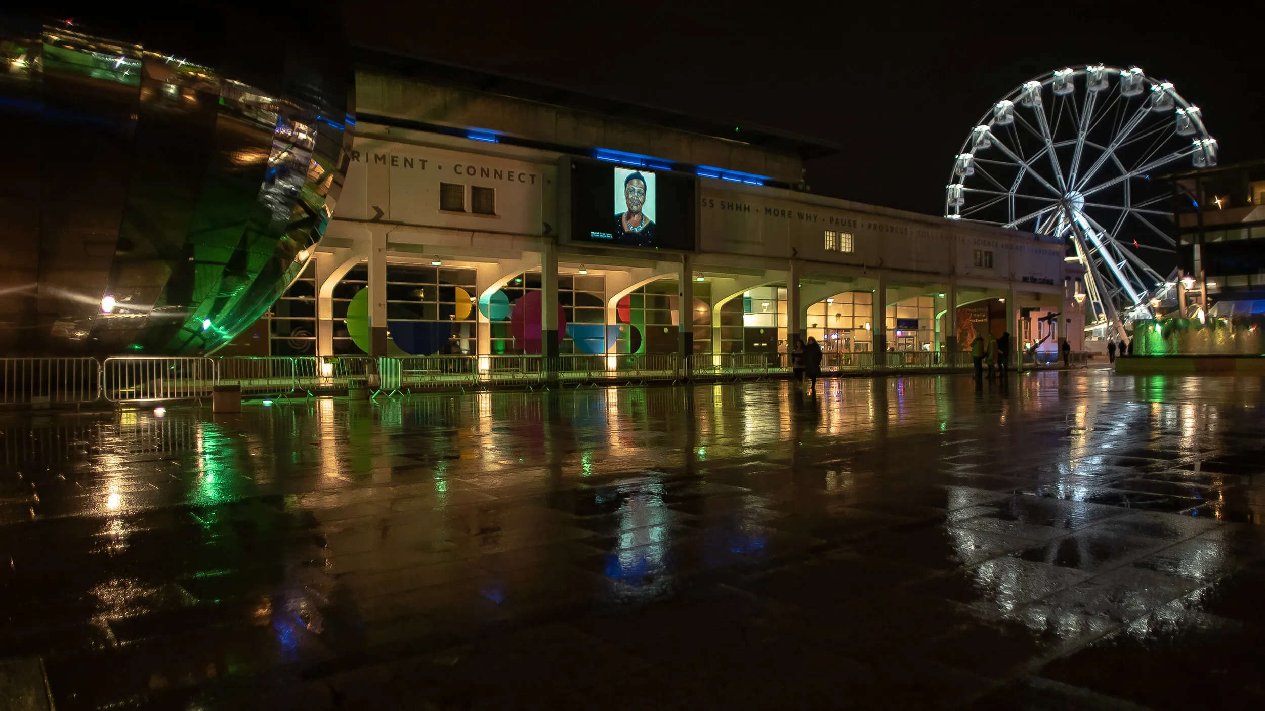 Night view of Millennium Square with a close-up painted portrait shown on the big screen, the illuminated observation wheel and We The Curious building reflected in the rain-soaked square