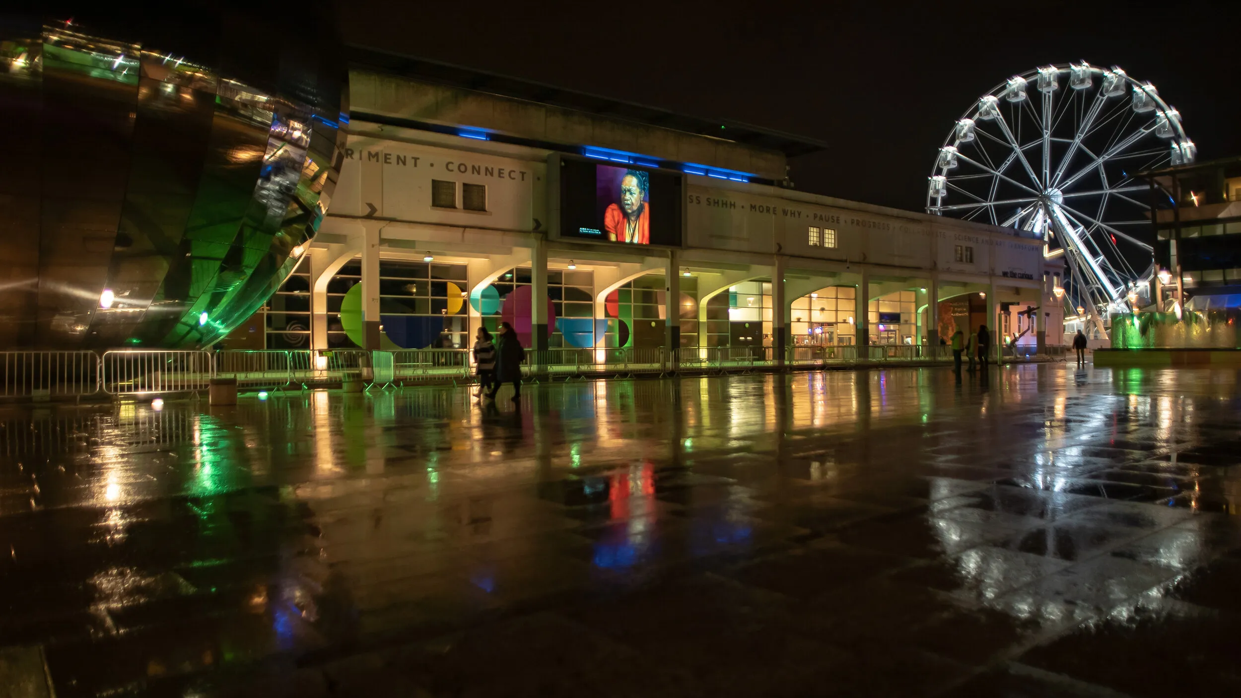 Night view of Millennium Square with a different painted portrait shown on the big screen, two visitors crossing the wet square