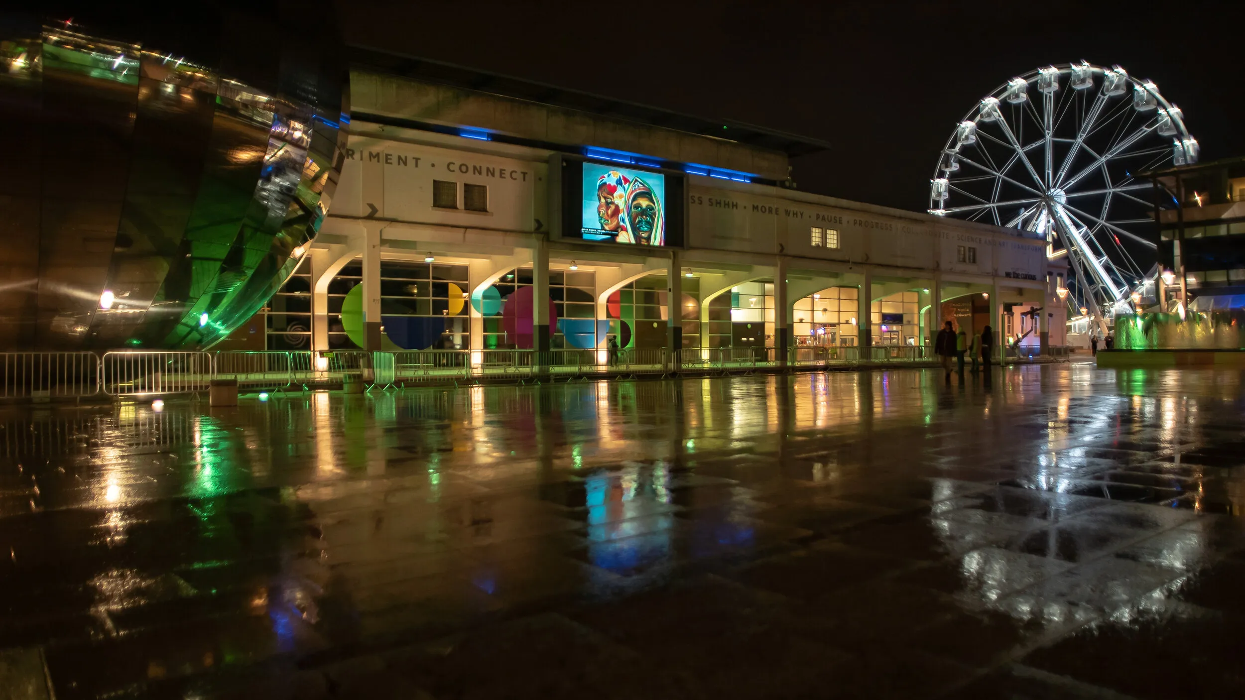 Night view of Millennium Square with a painted double portrait of two women in colourful headwraps shown on the big screen