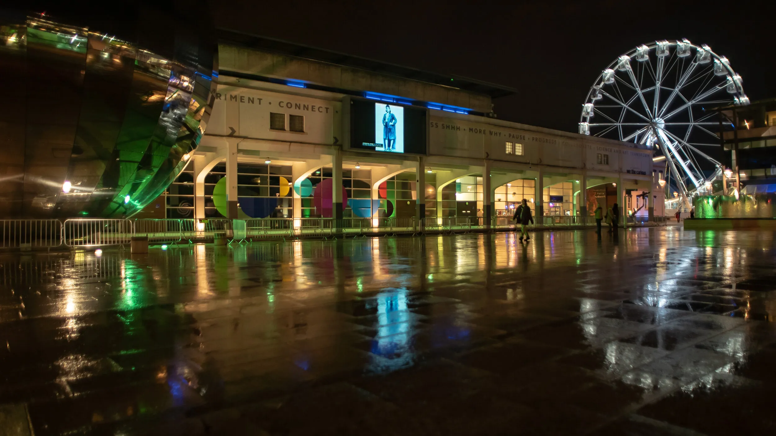 Night view of Millennium Square with a monochrome blue standing portrait shown on the big screen, a pedestrian crossing the wet square