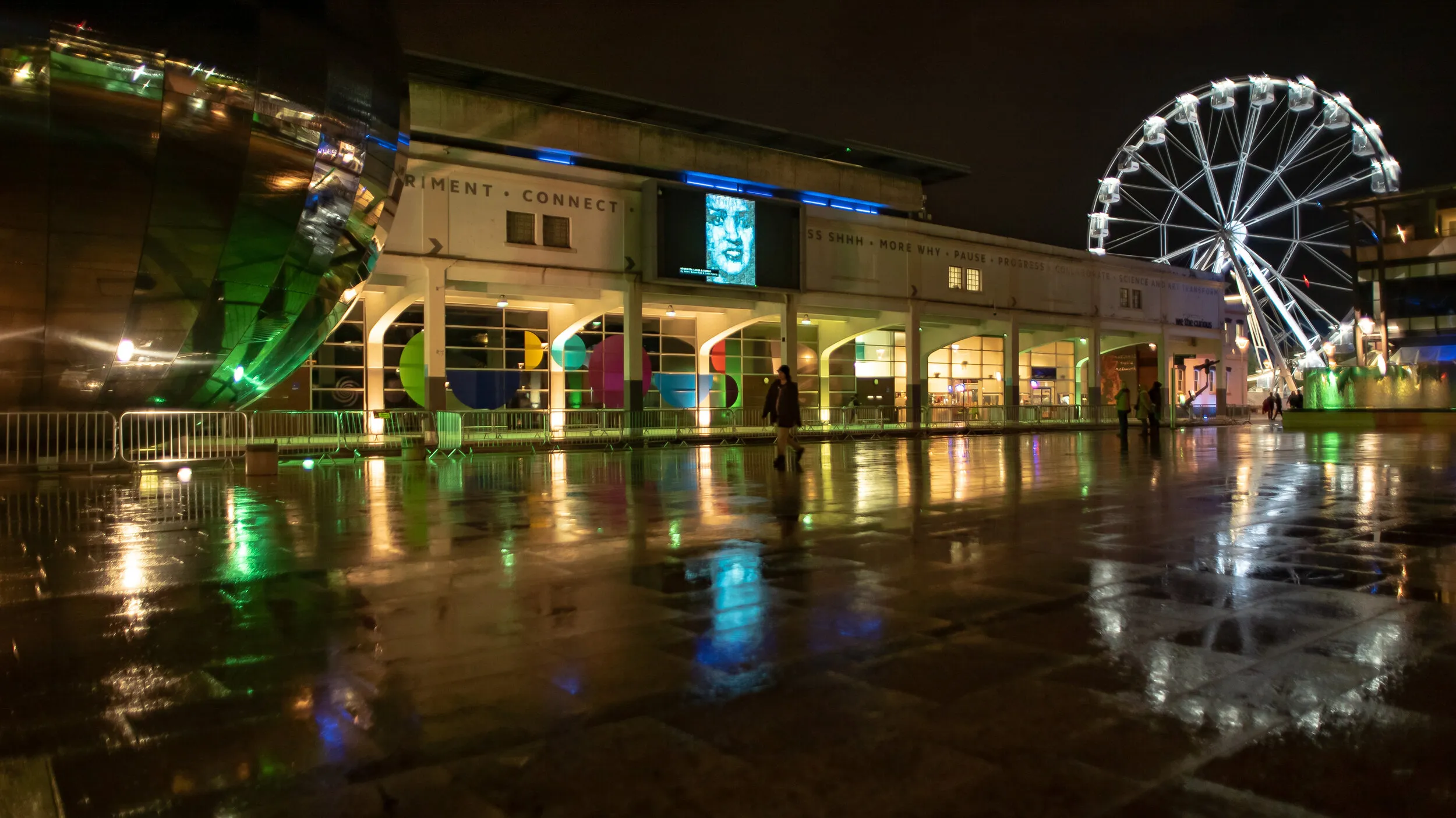 Night view of Millennium Square with a blue pixel-mosaic portrait shown on the big screen