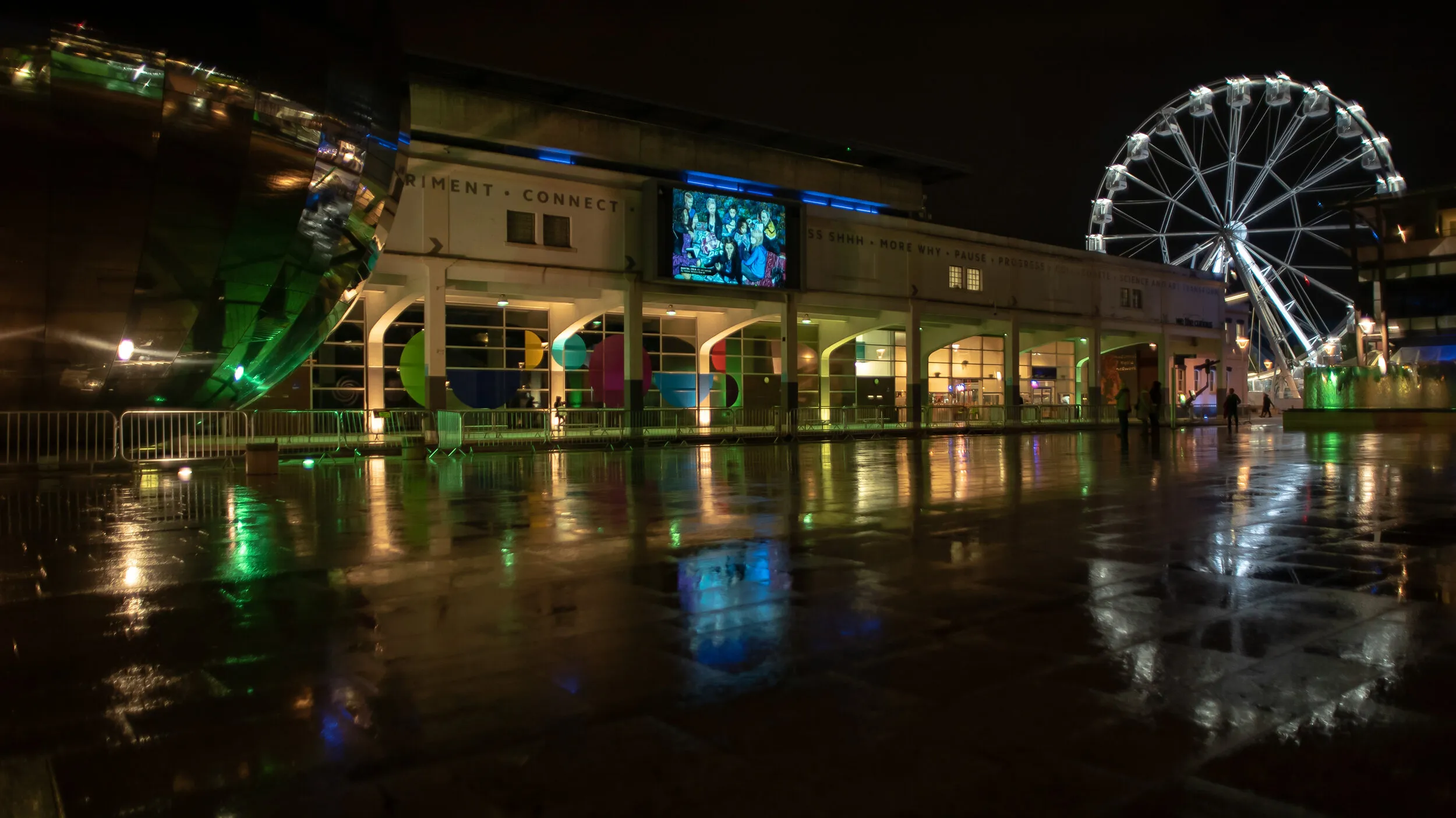 Night view of Millennium Square with an information screen headed 'Bristol', showing a portrait and a list of credits, on the big screen