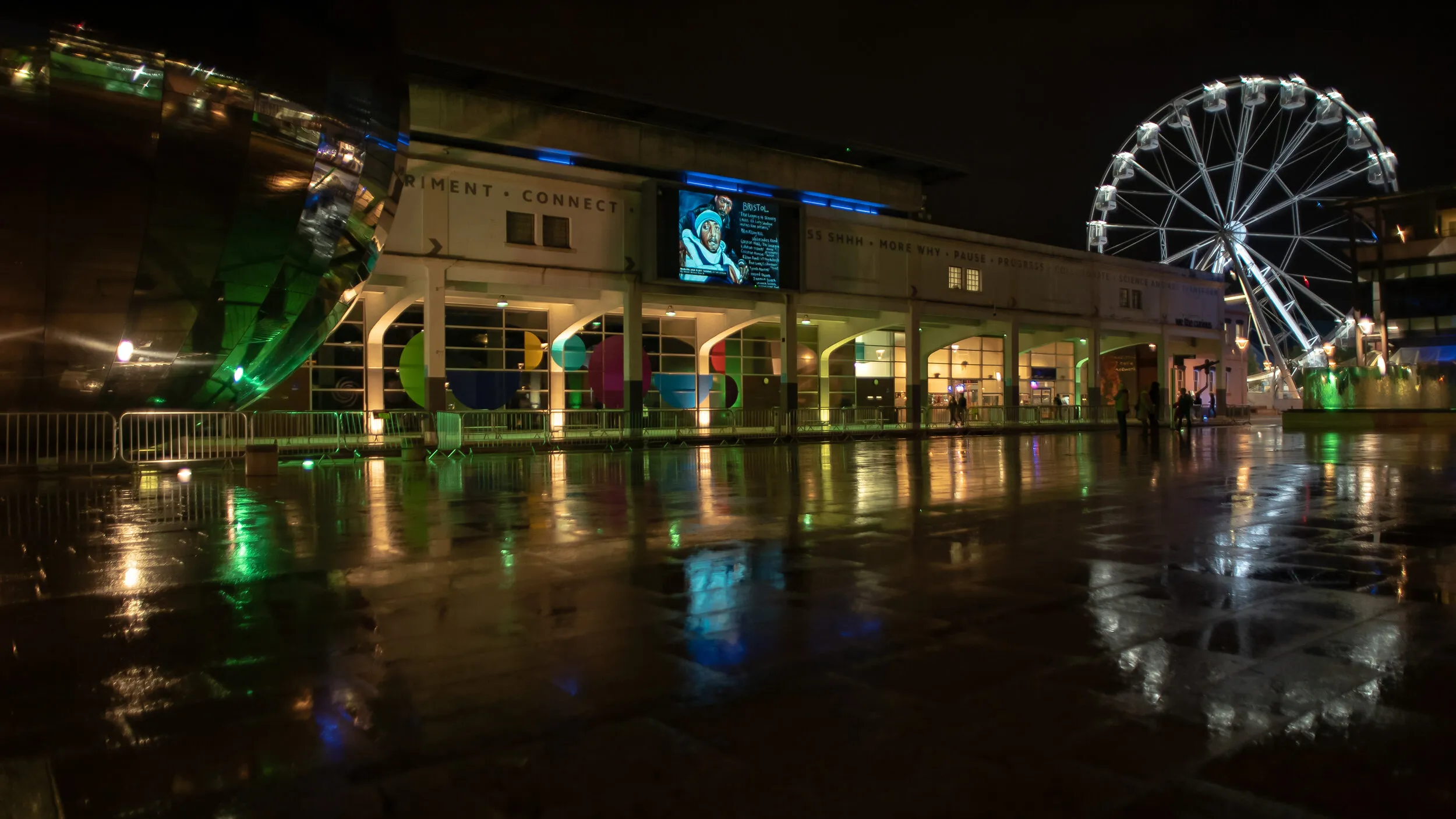 Night view of Millennium Square with a blue-toned portrait shown on the big screen, a cyclist crossing the wet square